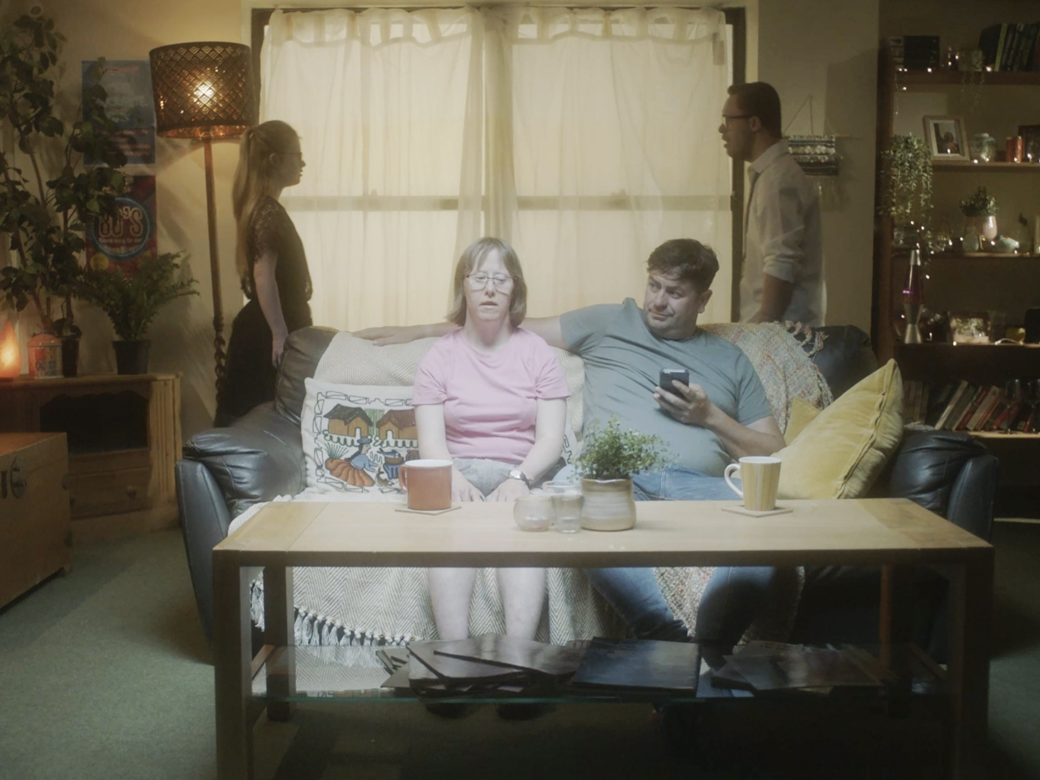 Production still from Typical? showing two people seated on a sofa in a living room while two figures stand in silhouette behind them.